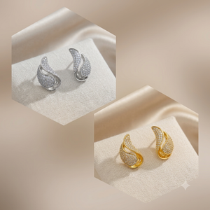 Two pairs of curved, pavé-set stud earrings displayed on a soft cream fabric surface. One pair is silver-toned and the other is gold-toned, each set with small clear stones.