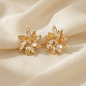 A pair of gold flower-shaped stud earrings with white inlay petals, displayed on a draped cream satin fabric background.