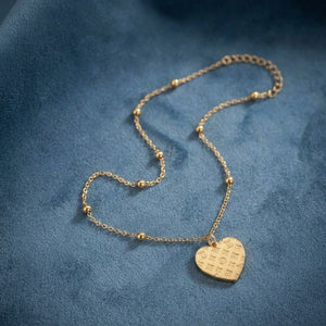 A delicate gold chain necklace with small bead accents and a heart-shaped pendant engraved with script, displayed on a textured blue fabric background.