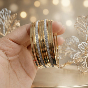 A hand holding a stack of slim bangles, including gold-toned solid bands and textured silver-toned bands, against a warm bokeh background with decorative gold elements.