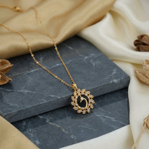 A gold necklace with a circular floral pendant set with small clear stones, displayed on a dark gray marble block with draped beige satin fabric and dried floral props.