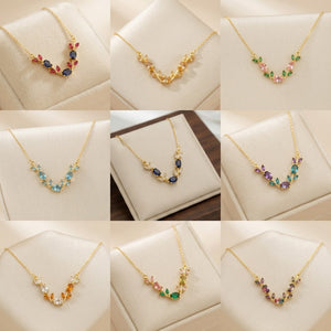 A 3x3 collage showing nine close-up views of gold-tone necklaces displayed on cream jewelry stands. Each necklace has a delicate leaf or vine motif with small multicolored gemstones in red, blue, green, pink, purple, orange, and clear stones.