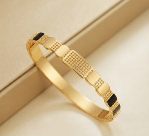 A gold-plated bangle bracelet with alternating smooth rectangular links and textured square links, accented with small black inlays, displayed on a beige surface.