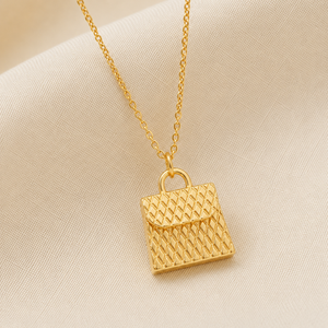 A gold necklace with a textured, diamond-patterned handbag pendant, displayed on a soft beige fabric background.