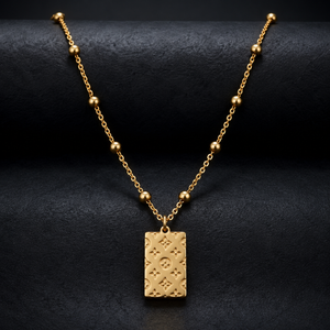 A gold necklace with a rectangular engraved pendant and beaded chain, displayed on a textured black background.