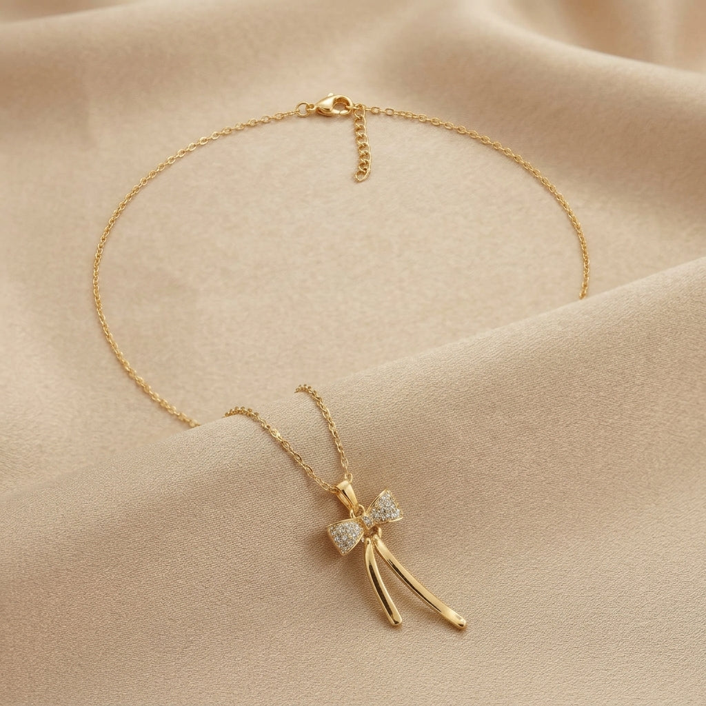 A delicate gold chain necklace with a bow pendant adorned with small clear stones, displayed on a soft beige fabric background.