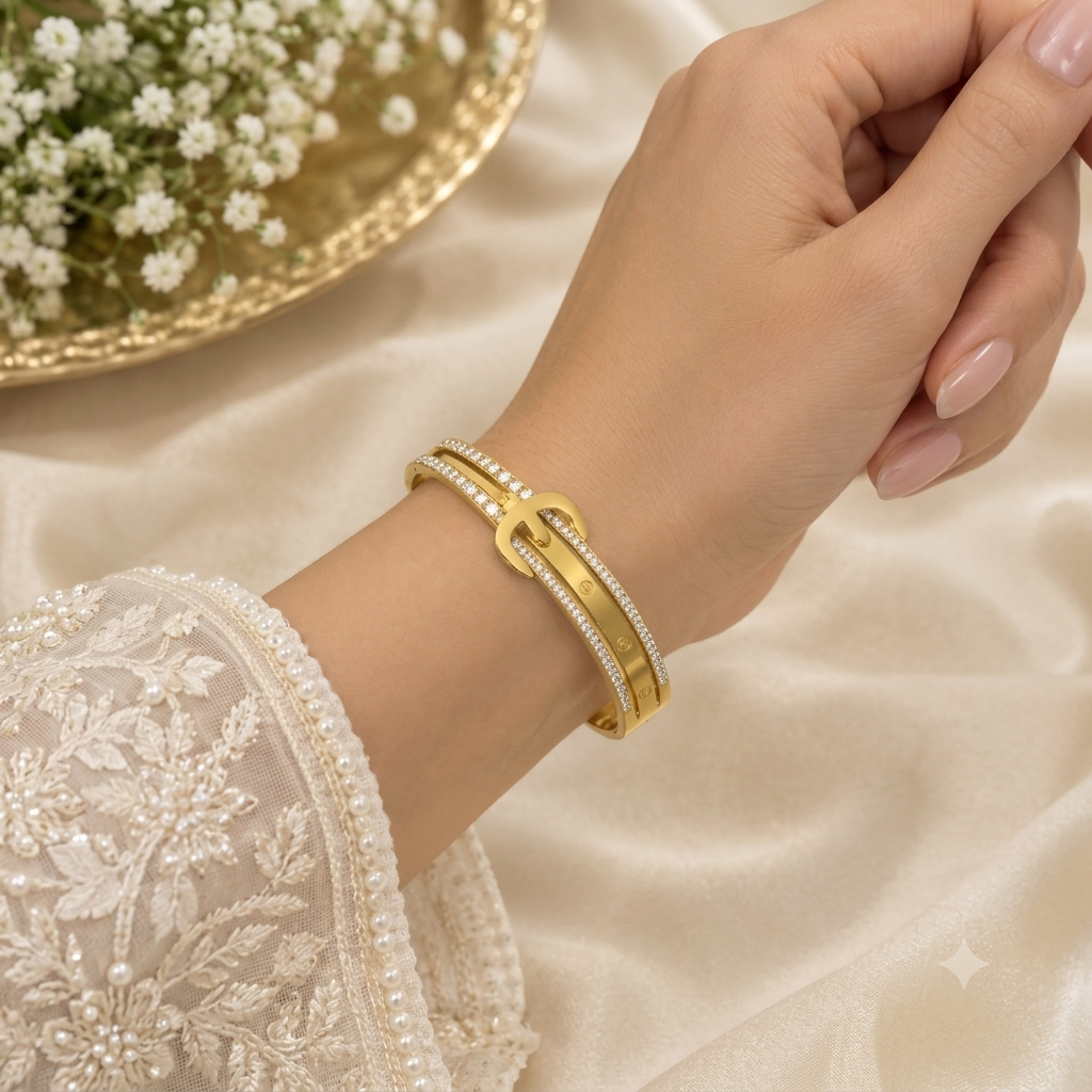 A close-up of a woman's wrist wearing a double-strand gold bangle bracelet with pavé-set clear stones and a central interlocking gold motif. The image is styled with a white lace sleeve, cream satin fabric, and a gold bowl of baby's breath in the background.