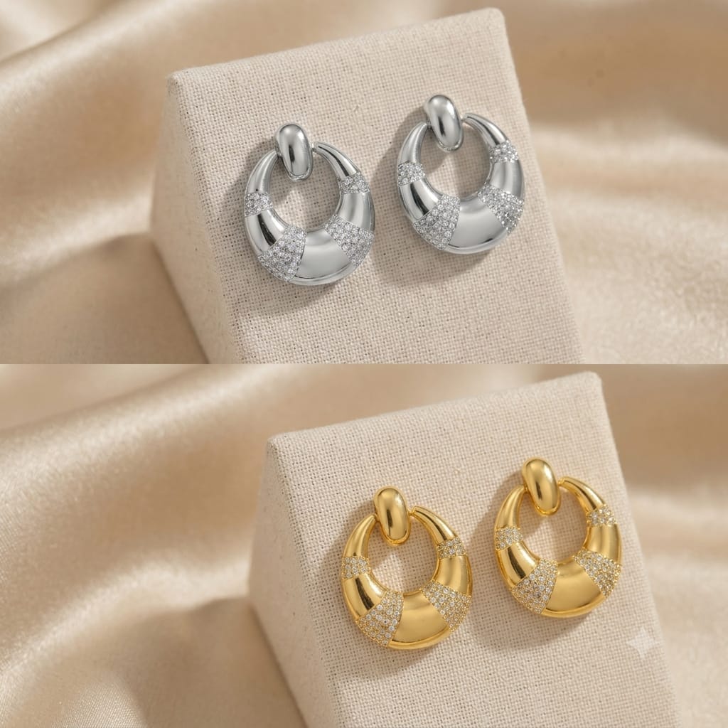 Two pairs of crescent-shaped hoop earrings displayed on beige fabric jewelry stands. The top pair is polished silver with pavé-set clear stones, and the bottom pair is polished yellow gold with the same stone detailing.