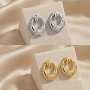 Two pairs of crescent-shaped hoop earrings displayed on beige fabric jewelry stands. The top pair is polished silver with pavé-set clear stones, and the bottom pair is polished yellow gold with the same stone detailing.