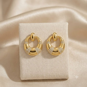 A pair of gold crescent-shaped hoop earrings with pavé-set clear stones, displayed on a textured beige jewelry stand against a draped beige satin background.