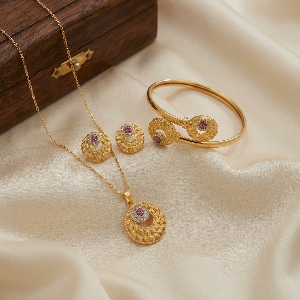Gold jewelry set featuring a necklace, bangle, and earrings with circular textured pendants and matching round studs, each accented with a small pink stone, displayed on cream satin fabric next to a dark wooden box.