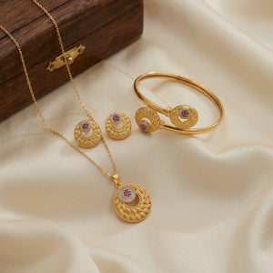 Gold jewelry set featuring a necklace, bangle, and earrings with circular textured pendants and matching round studs, each accented with a small pink stone, displayed on cream satin fabric next to a dark wooden box.