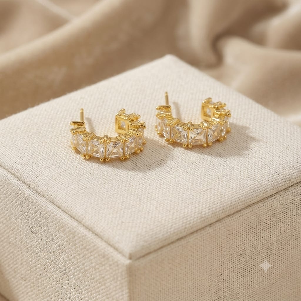 A pair of gold crescent-shaped hoop earrings with clear rectangular crystals, displayed on a textured beige jewelry box with a soft beige fabric background.