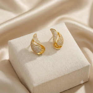 A pair of gold crescent-shaped earrings covered in small clear stones, displayed on a cream fabric jewelry box against a draped satin background.