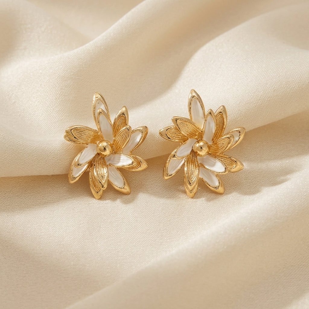 A pair of gold flower-shaped stud earrings with white inlay petals, displayed on a draped cream satin fabric background.