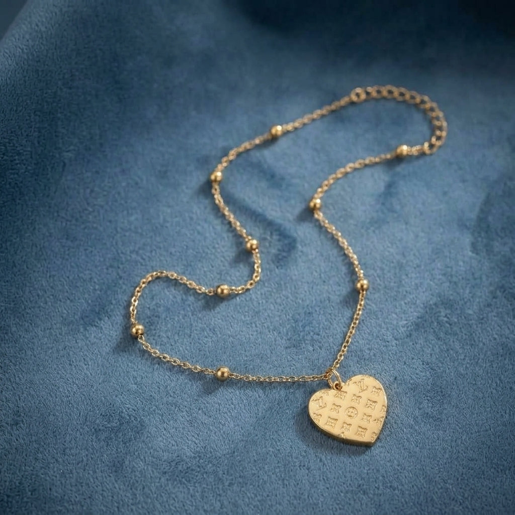 A delicate gold chain necklace with small bead accents and a heart-shaped pendant engraved with script, displayed on a textured blue fabric background.