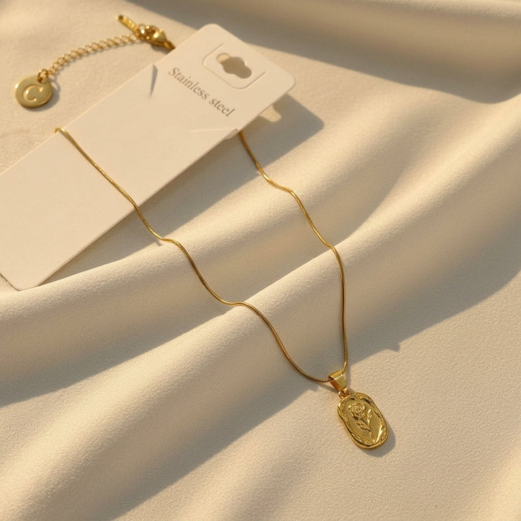 A gold-toned stainless steel necklace with a small oval pendant featuring a raised chevron design, displayed on a cream fabric background with its white hangtag.