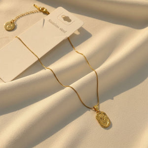 A gold-toned stainless steel necklace with a small oval pendant featuring a raised chevron design, displayed on a cream fabric background with its white hangtag.