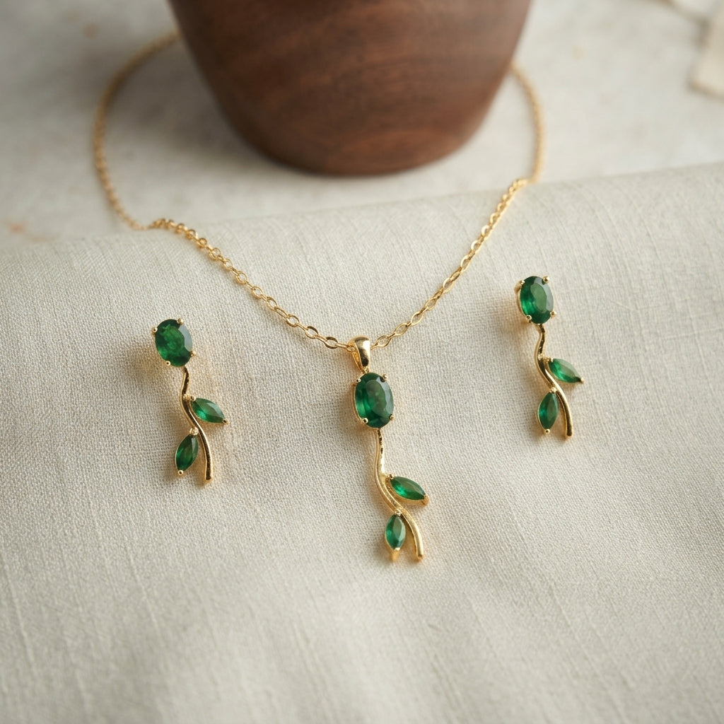 Gold jewelry set featuring a necklace and matching earrings with green gemstone leaf motifs, displayed on a textured cream fabric with a wooden bowl in the background.
