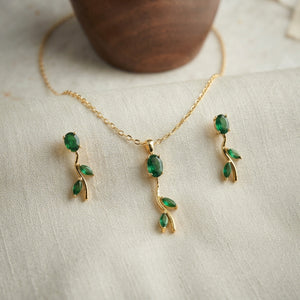 Gold jewelry set featuring a necklace and matching earrings with green gemstone leaf motifs, displayed on a textured cream fabric with a wooden bowl in the background.