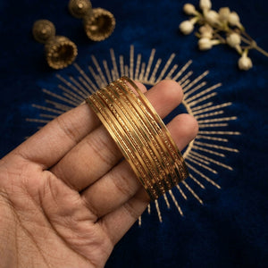 A hand holding a stack of gold-toned bangles against a dark blue velvet background with decorative gold and white floral elements.