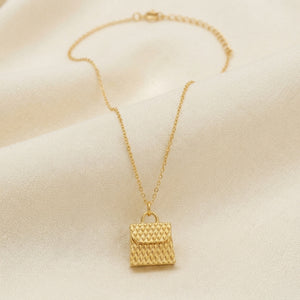 A delicate gold chain necklace featuring a small textured pouch-shaped pendant, displayed on a soft, draped cream-colored fabric background.