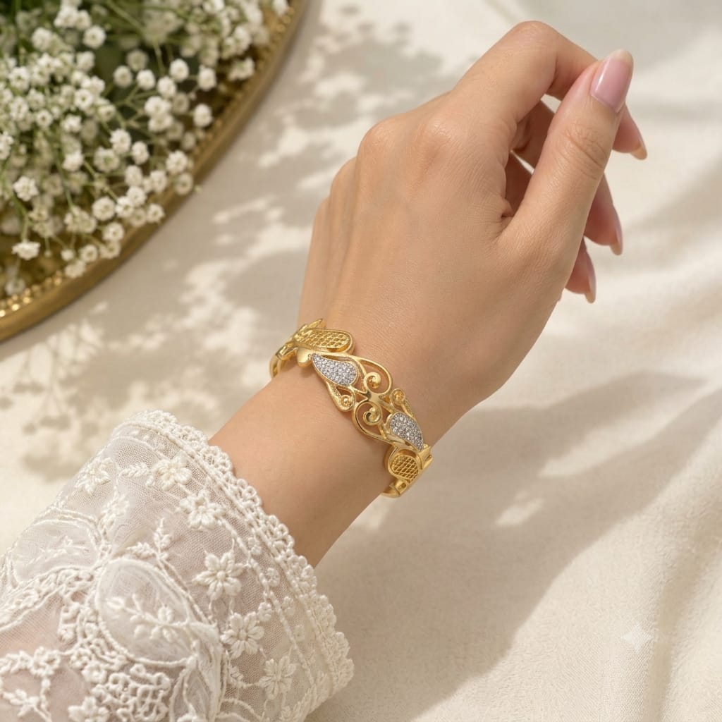 A gold-toned ornate bracelet with pavé-set clear stones, shown worn on a woman's wrist. The hand has light pink manicured nails, and the person is wearing a white lace long-sleeve blouse.