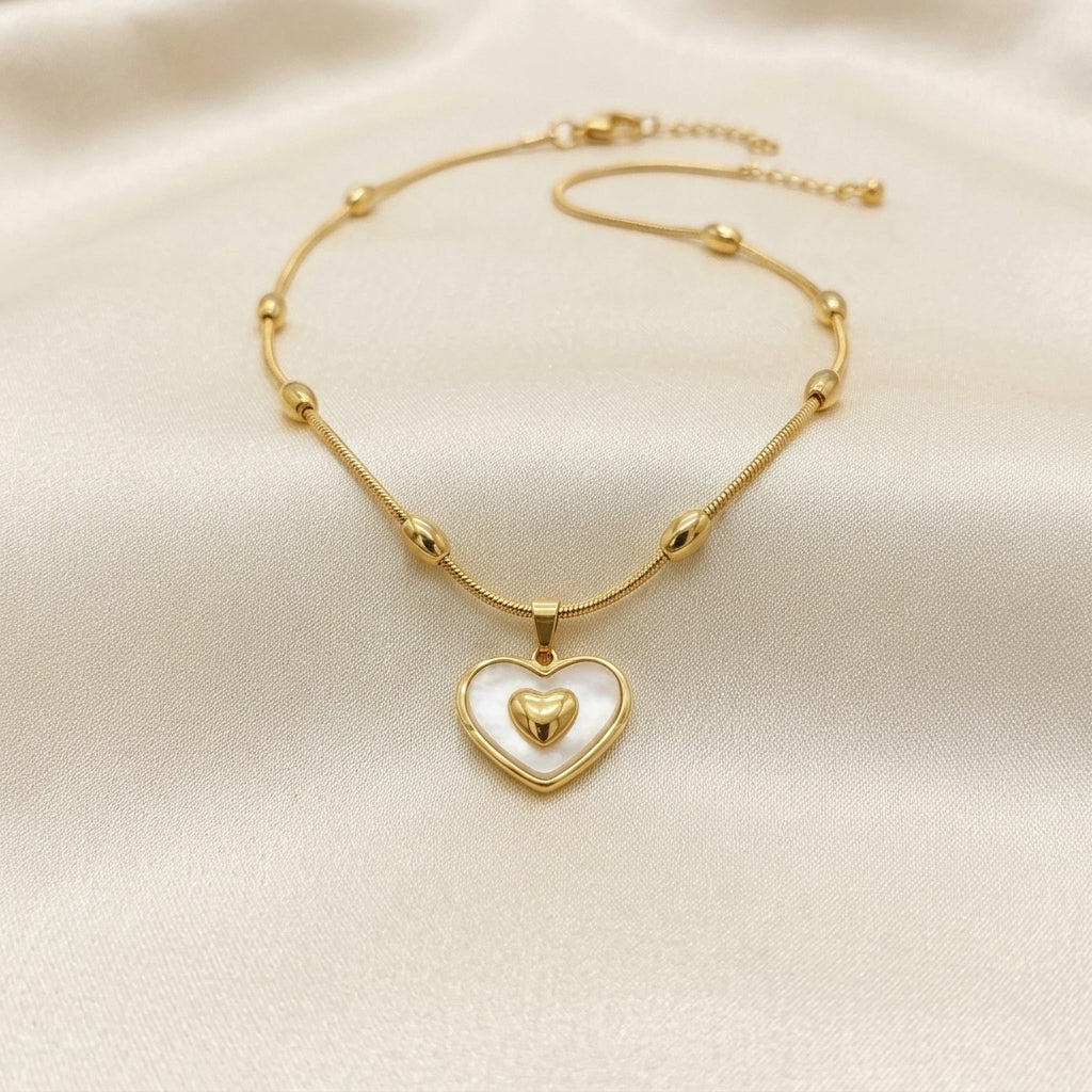 A delicate gold chain necklace with small bead accents and a heart-shaped pendant featuring a white inlay and a smaller gold heart in the center, displayed on a cream satin fabric background.