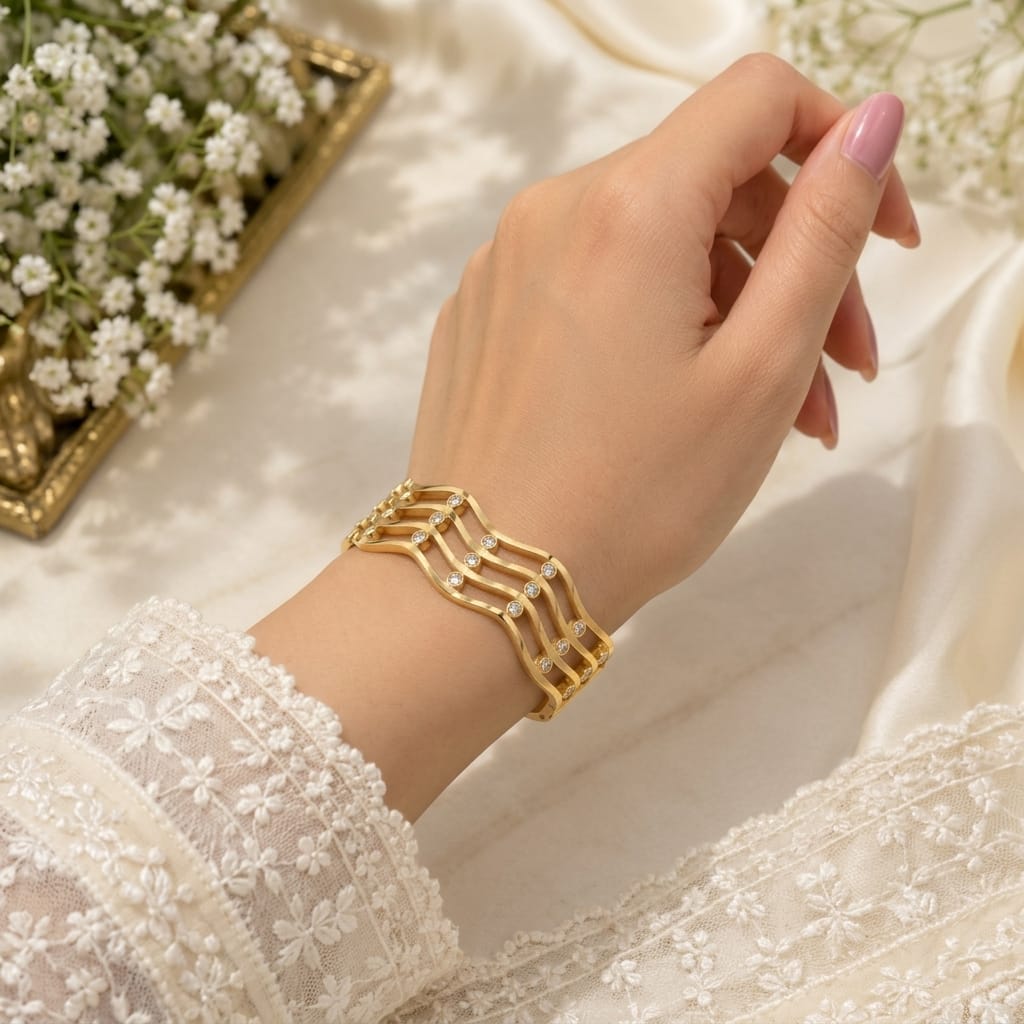 A gold-toned multi-strand geometric cuff bracelet with small clear rhinestones, worn on a woman's wrist. The hand has light pink manicured nails, and the person is wearing a white lace long-sleeve top.