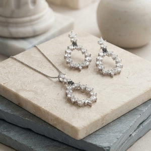 Silver-tone jewelry set with a necklace and matching drop earrings, each featuring a halo of clear cubic zirconia stones with a small black stone accent, displayed on stacked stone blocks.