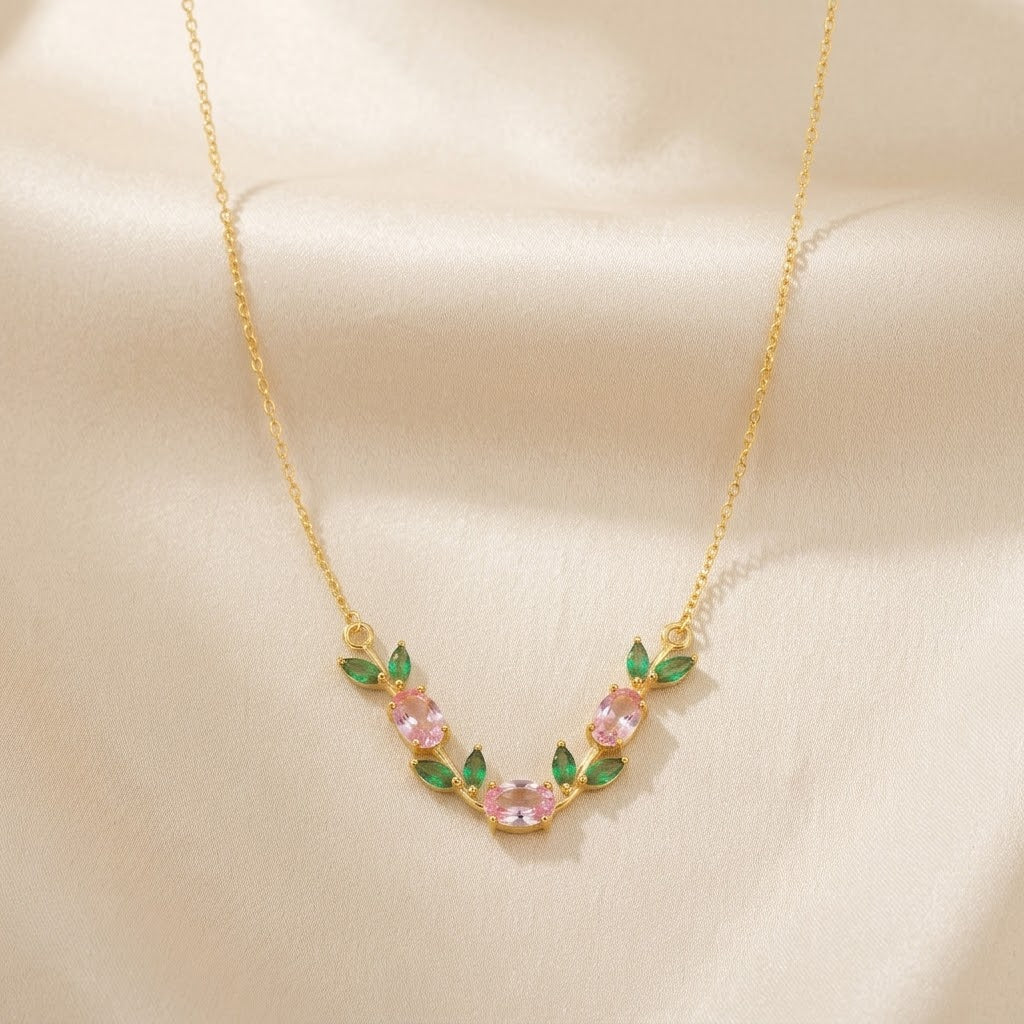 A delicate gold necklace featuring a curved floral design with pink marquise-cut stones and green leaf accents, displayed on a soft beige satin fabric background.