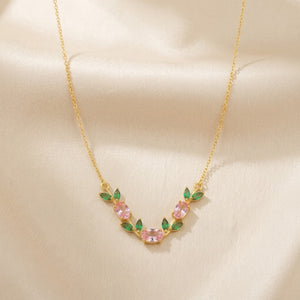 A delicate gold necklace featuring a curved floral design with pink marquise-cut stones and green leaf accents, displayed on a soft beige satin fabric background.