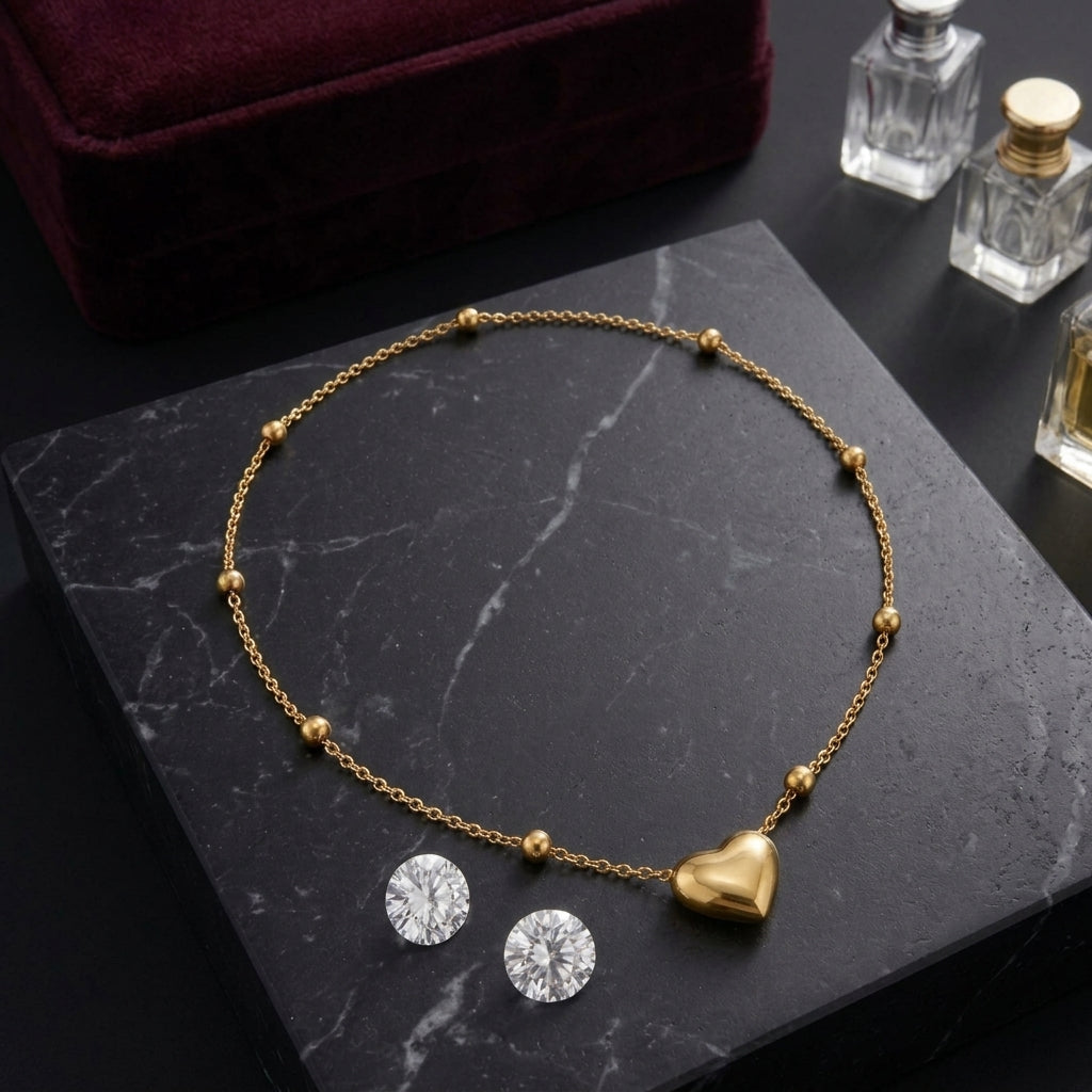 A gold chain necklace with small bead accents and a polished gold heart pendant, displayed on a black marble surface next to two round clear gemstones. A dark burgundy velvet jewelry box is partially visible in the background, along with small glass perfume bottles.