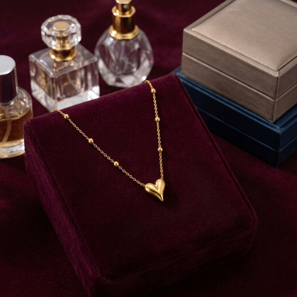 A gold heart pendant necklace draped over a deep burgundy velvet jewelry box, with perfume bottles and stacked gift boxes in the background.