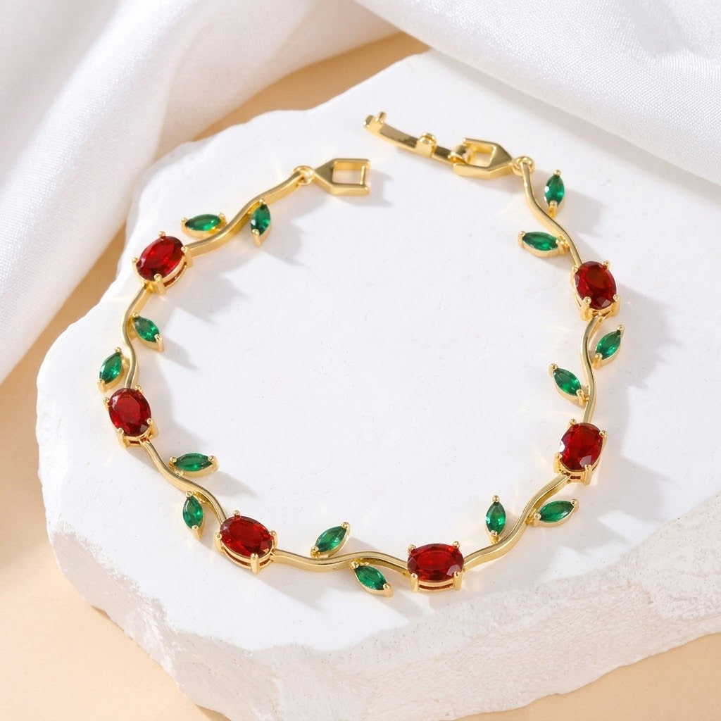 A gold vine-style bracelet with red oval gemstones and green leaf accents, displayed on a white stone slab with draped white fabric in the background.