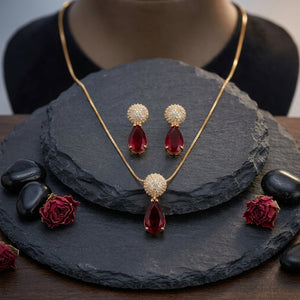 Gold jewelry set with a necklace and matching earrings, featuring deep red teardrop gemstones and round crystal-encrusted tops, displayed on a dark slate stand with decorative red roses and black stones.