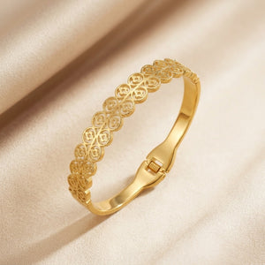 A gold bangle bracelet with an intricate openwork filigree pattern, resting on a draped beige satin fabric background.