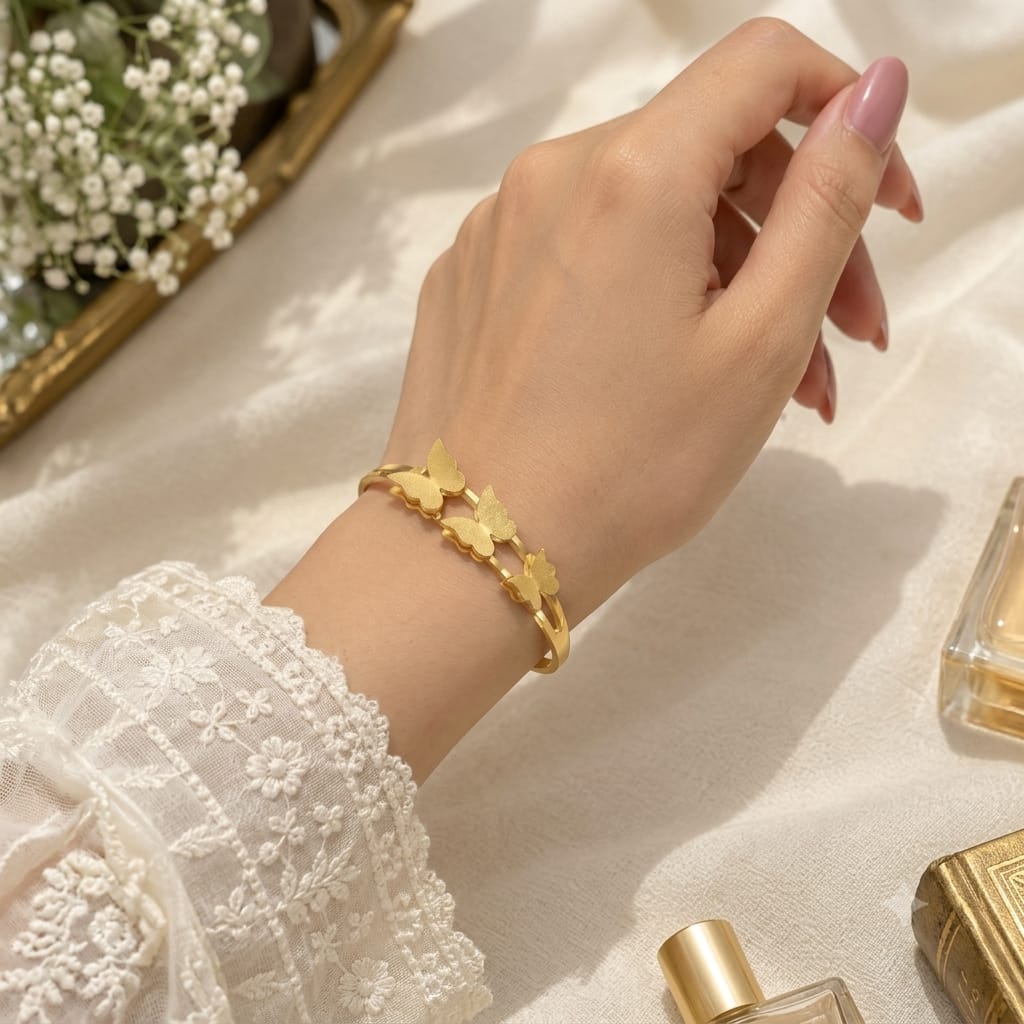 A gold butterfly bracelet worn on a woman's wrist, styled with a white lace sleeve against a soft cream fabric background with decorative flowers and perfume bottles.