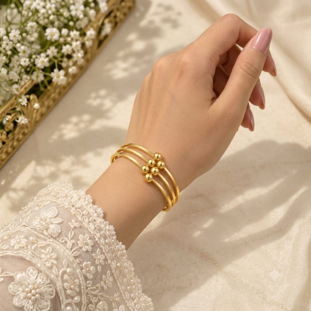 A close-up of a person's hand and wrist wearing a gold triple-band bracelet with a central floral bead design, styled with a white lace sleeve against a soft beige fabric background.