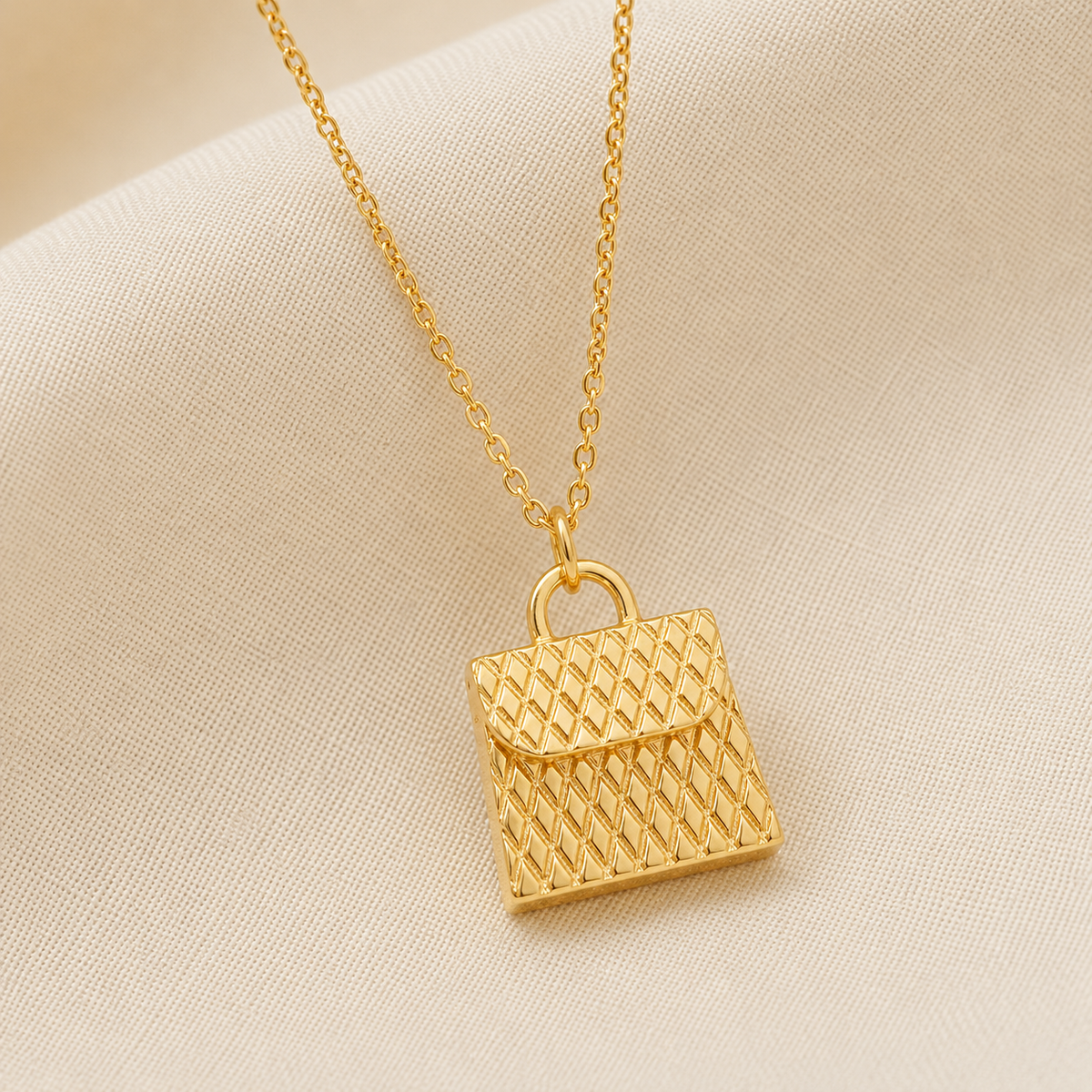 A gold necklace with a textured, diamond-patterned handbag pendant, displayed on a soft beige fabric background.