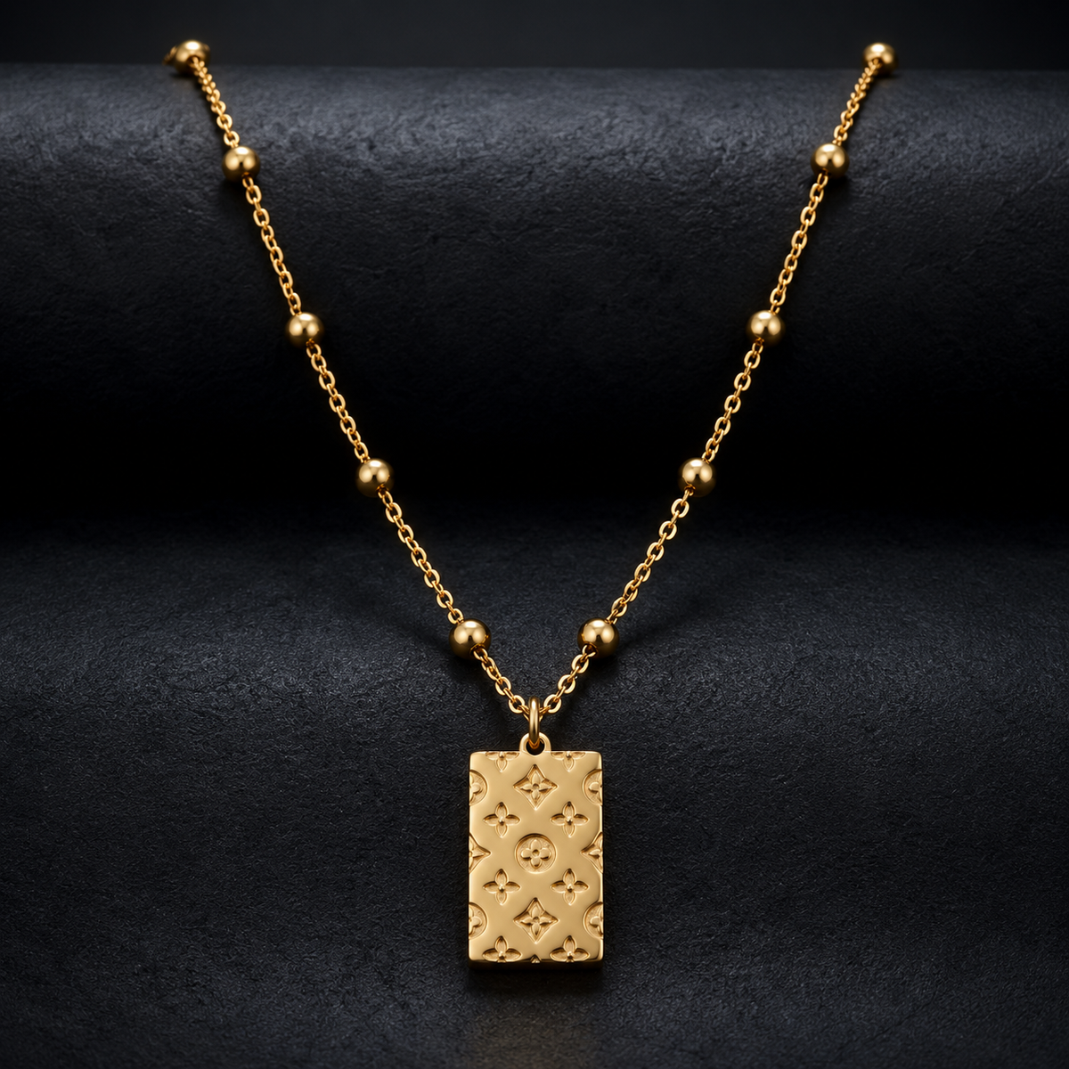 A gold necklace with a rectangular engraved pendant and beaded chain, displayed on a textured black background.